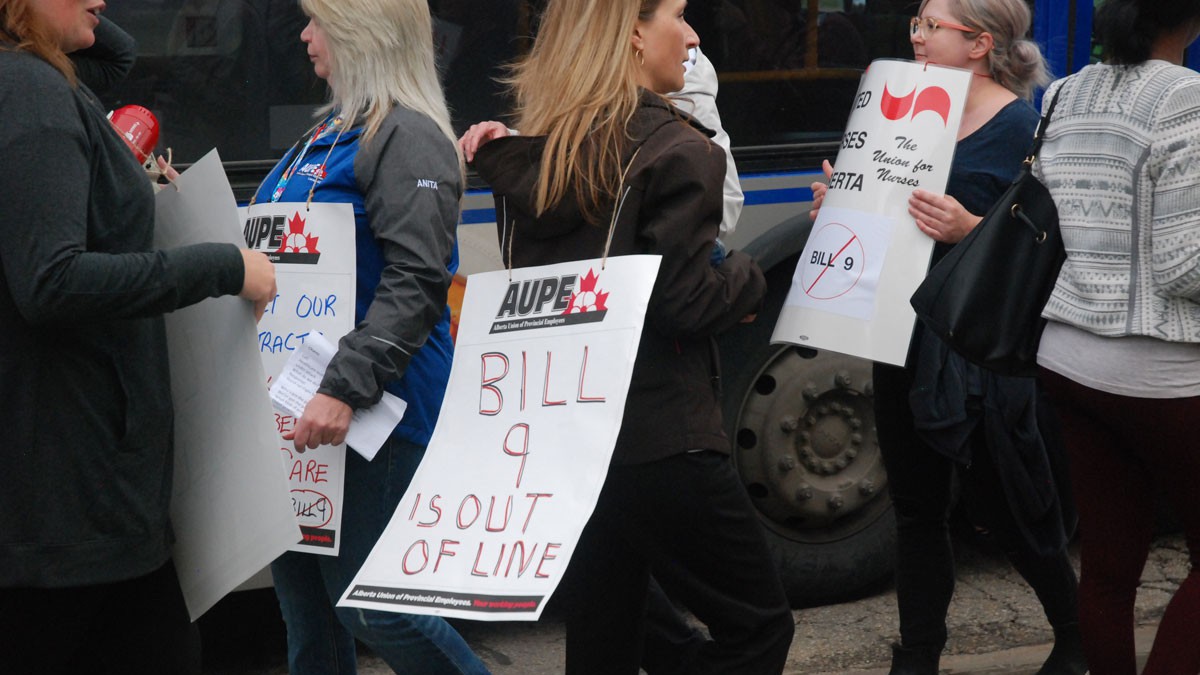 A person with long hair walks a picket line among others, wearing a sign that says "Bill 9 is out of line."